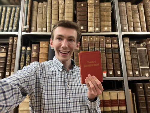 Young man in library holding a red book in front of old bookshelves.