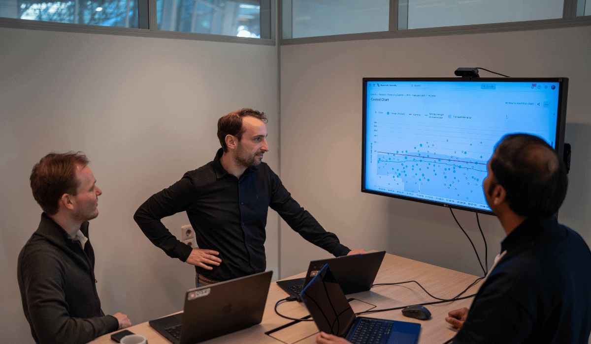 Three men in a meeting room discussing data on a large screen.
