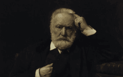A man with white hair and beard, dressed in dark clothing, sitting in a contemplative pose.