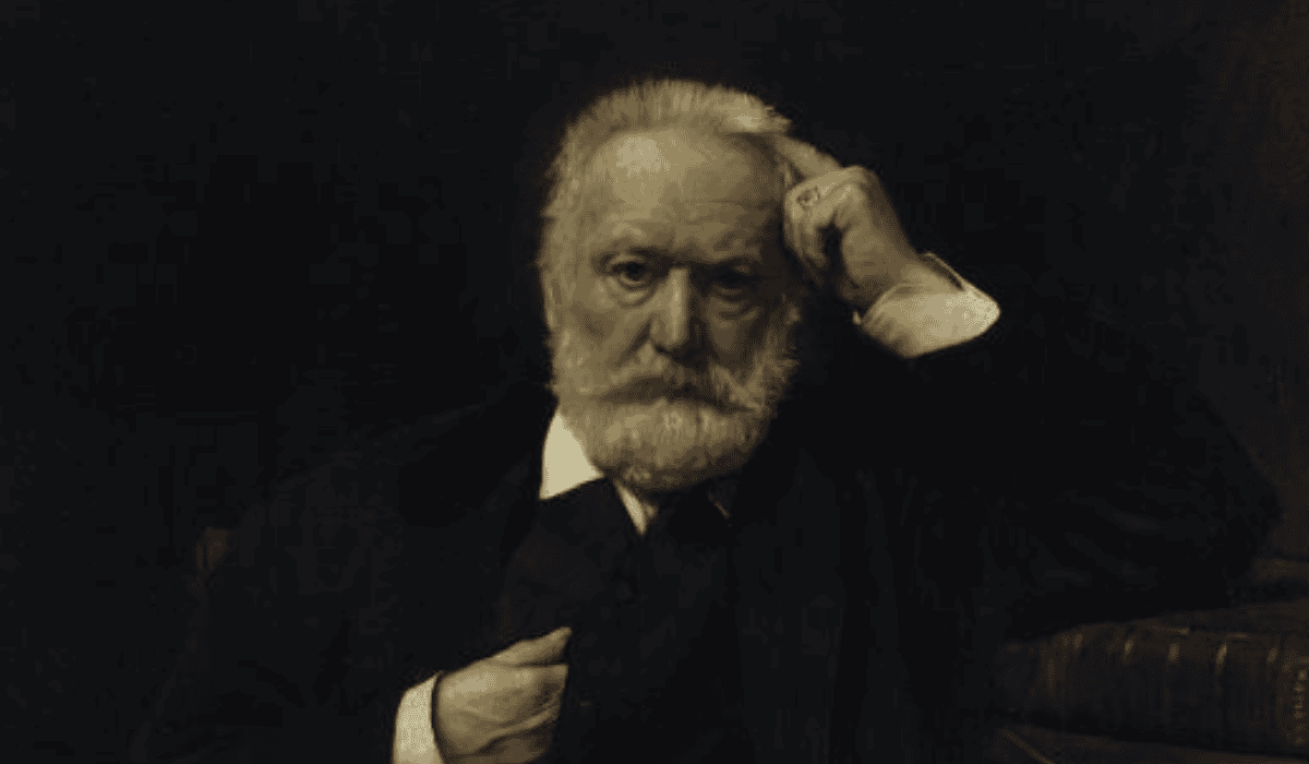 A man with white hair and beard, dressed in dark clothing, sitting in a contemplative pose.